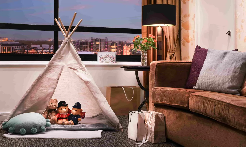 Elegant hotel room at Millennium Knightsbridge featuring a cozy teepee play tent with plush toys, a brown sofa, decorative cushions, and a city view through large windows.