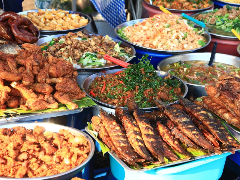 Assorted cooked dishes including fried chicken, grilled fish, stir-fried vegetables, and noodles served in metal bowls and trays at a street food market.