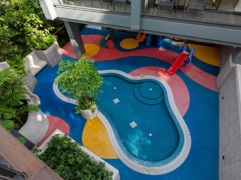 Outdoor area with a small, irregularly-shaped swimming pool surrounded by colorful flooring, a children's slide into the pool, potted plants, and a partially covered upper level with glass railings.
