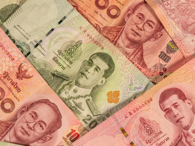 Scattered Thai banknotes in denominations of 20, 50, and 100 Baht, featuring traditional designs and historical figures, partially obscured by rectangular blocks.