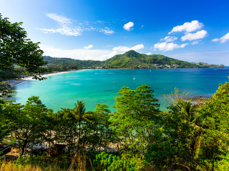 A picturesque coastal scene featuring clear turquoise waters, verdant vegetation, and distant hills beneath a bright blue sky. The tranquil setting is accentuated by boats gently floating in the bay, embodying the serene beauty of nature.