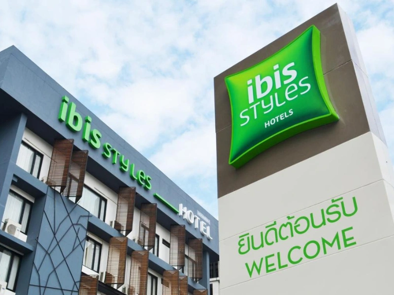 Exterior view of an Ibis Styles hotel featuring two signs: one on the building reading 'ibis STYLES HOTEL' in green, and a standalone sign with a green pillow-shaped logo and bilingual text in Thai and English saying 'ยินดีต้อนรับ WELCOME'.
