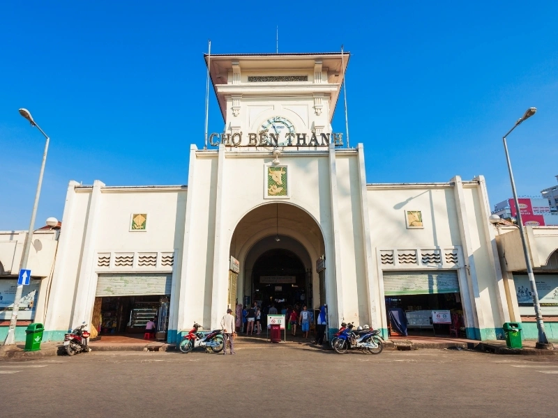 Front entrance of Bến Thành Market in Ho Chi Minh City, Vietnam, featuring a central clock tower with 'CHO BEN THANH' signage, motorbikes parked in front, and people entering and exiting on a sunny day.