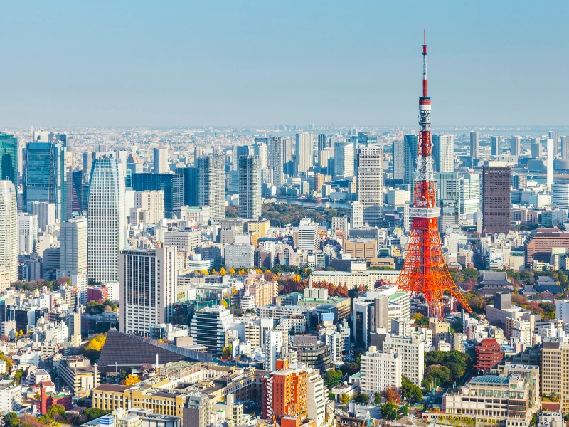 Panoramic view of Tokyo city with high-rise buildings and Tokyo Tower in the center-right, under a clear blue sky.
