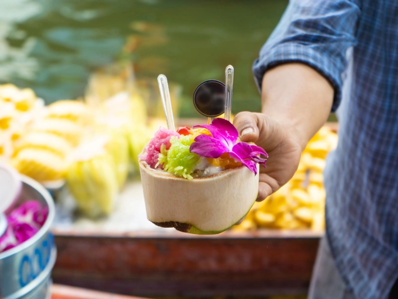 A person holding a coconut shell filled with colorful shaved ice or dessert. The dessert is topped with a purple orchid flower and has two spoons inserted into it. In the background, there are blurred images of yellow fruits, possibly bananas or pineapples, on a boat.