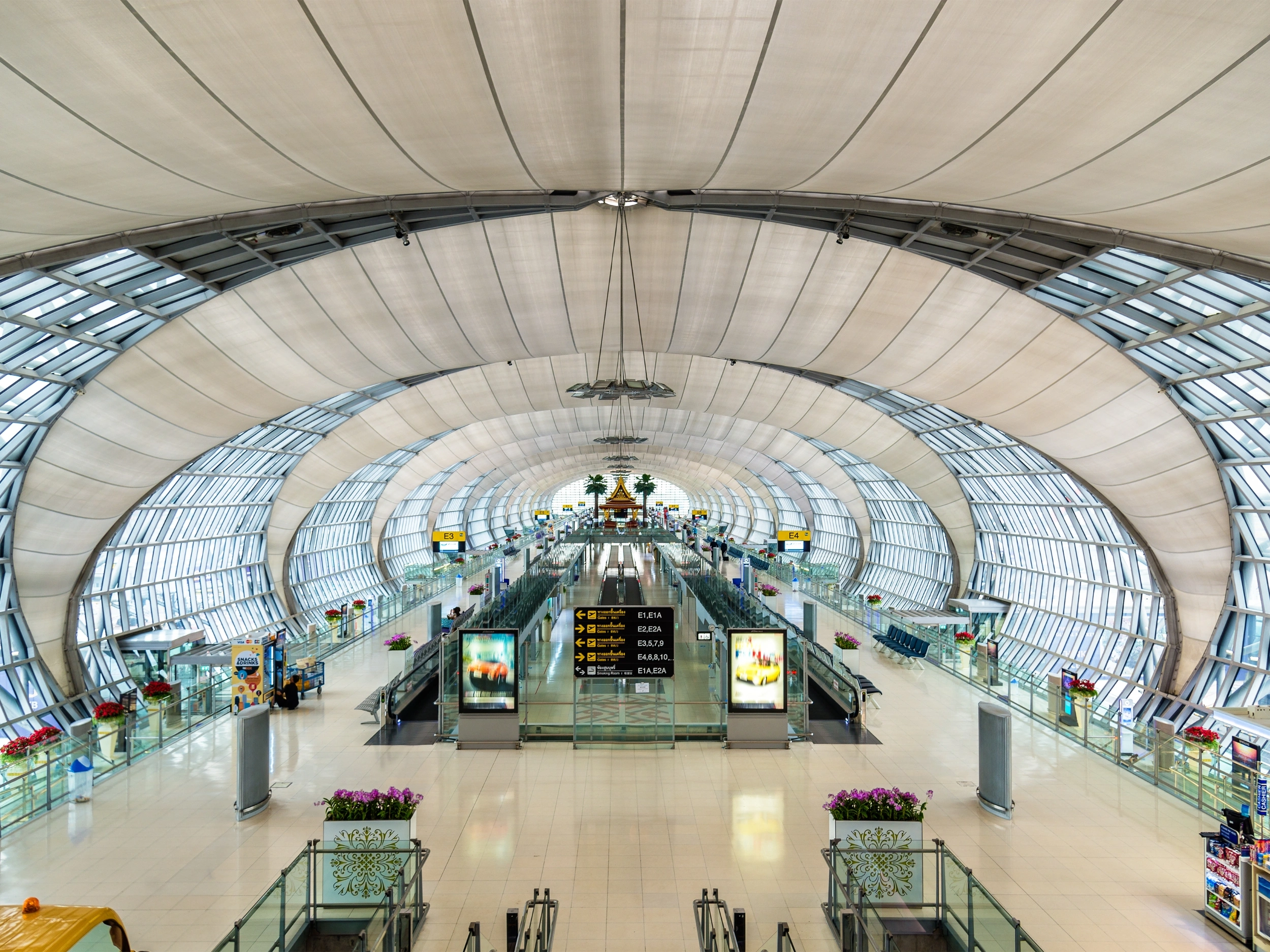 The interior of a modern airport terminal with a high, curved ceiling and large windows allowing natural light to enter. There are multiple escalators and walkways leading to different gates. In the center, there is an information board displaying flight details. The terminal is spacious and well-lit, with various signs and advertisements visible throughout.