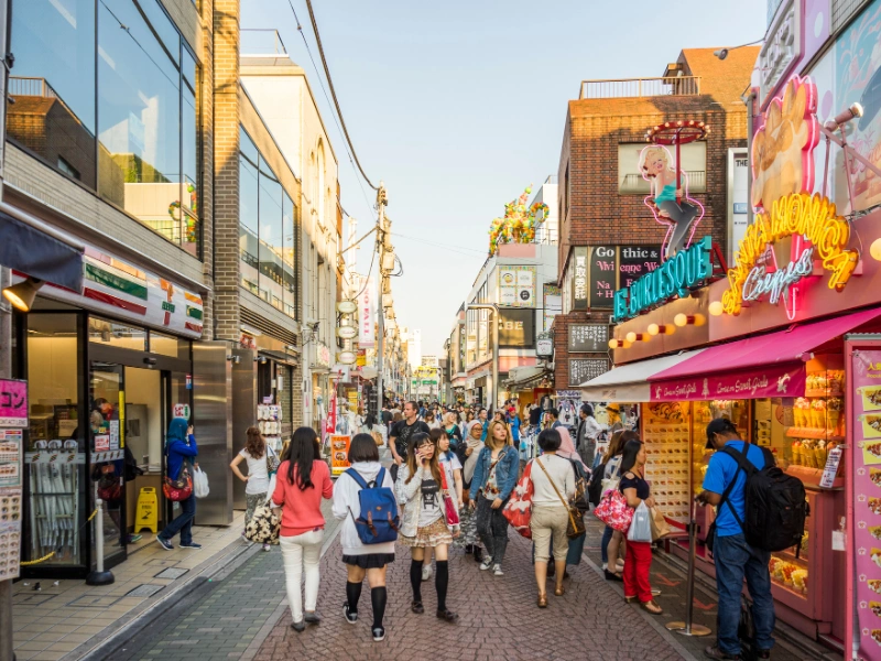 Crowded Takeshita Street in Harajuku, Tokyo, lined with vibrant shops including a 7-Eleven and a colorful crepe store, with people walking and shopping under bright signage in English and Japanese.