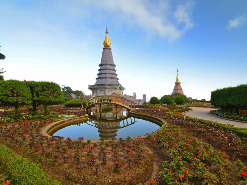 A landscaped garden with a pond, arched wooden bridge, and two golden-spired pagodas under a clear blue sky.