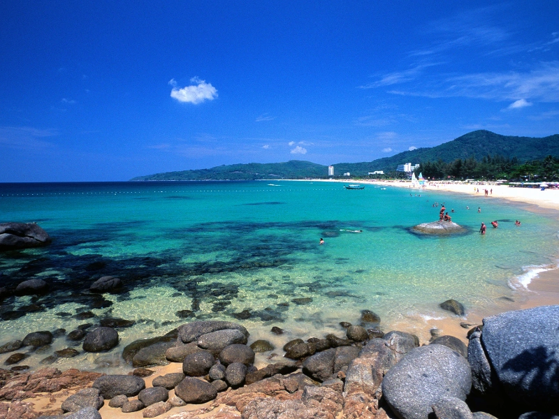 A scenic beach with turquoise waters, rocky foreground, sandy shore, and people swimming and relaxing. Green hills and a blue sky with scattered clouds complete the backdrop.