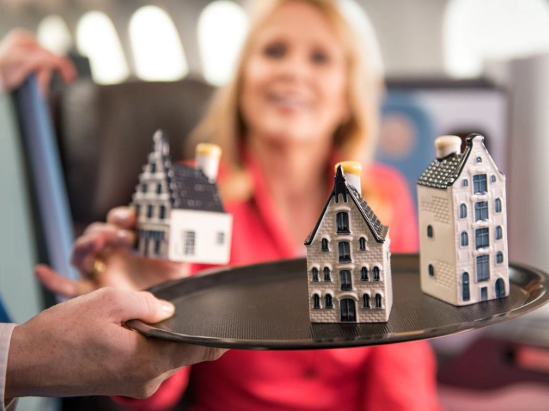 A tray holding three miniature ceramic houses with detailed facades, resembling traditional Dutch architecture. The houses are white with dark roofs and windows, and they are being presented by a person’s hand in an airplane cabin setting.