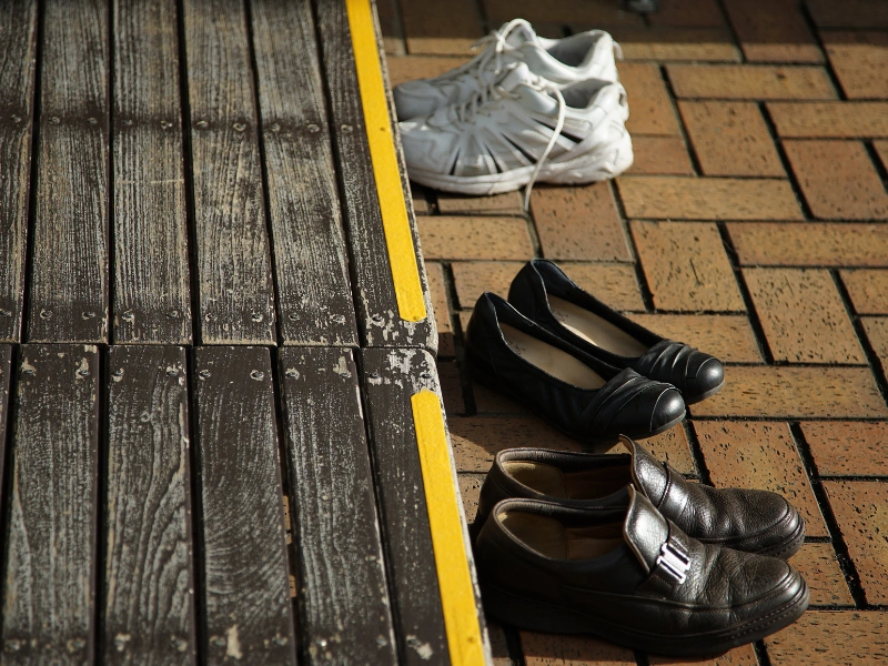 Three pairs of shoes—white sneakers, black flats, and brown loafers—placed on a brick floor beside a wooden deck with a yellow edge line, suggesting a no-shoes area.