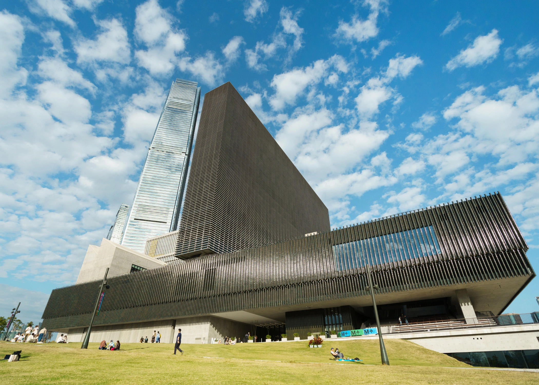 Exterior view of the M+ Museum in Hong Kong, showcasing its sleek, modern architecture with a horizontal podium and vertical tower, set against the Victoria Harbour waterfront. The facade features embedded LED panels used for displaying moving image works, visible from across the harbor