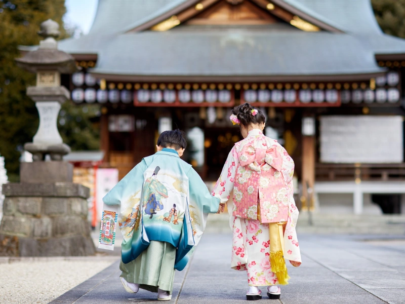 Two children wearing traditional kimonos—one in light blue with colorful patterns, the other in pink with floral designs—walking hand in hand toward a Japanese shrine with wooden structures and lanterns