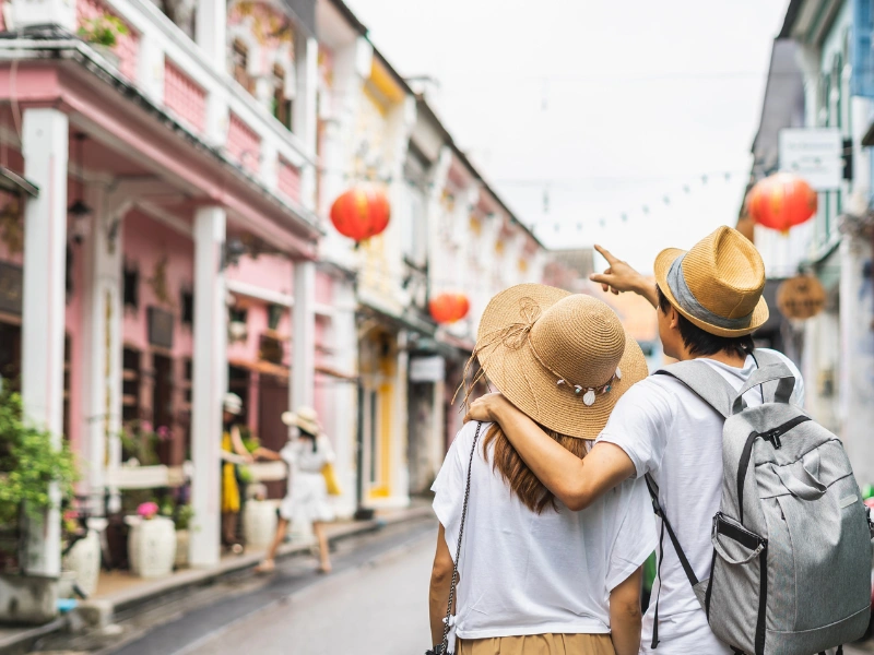 The image shows two people walking down a street with colorful buildings and red lanterns hanging above. The person on the right is pointing towards something in the distance. Both individuals are wearing hats and casual clothing, with one carrying a backpack.