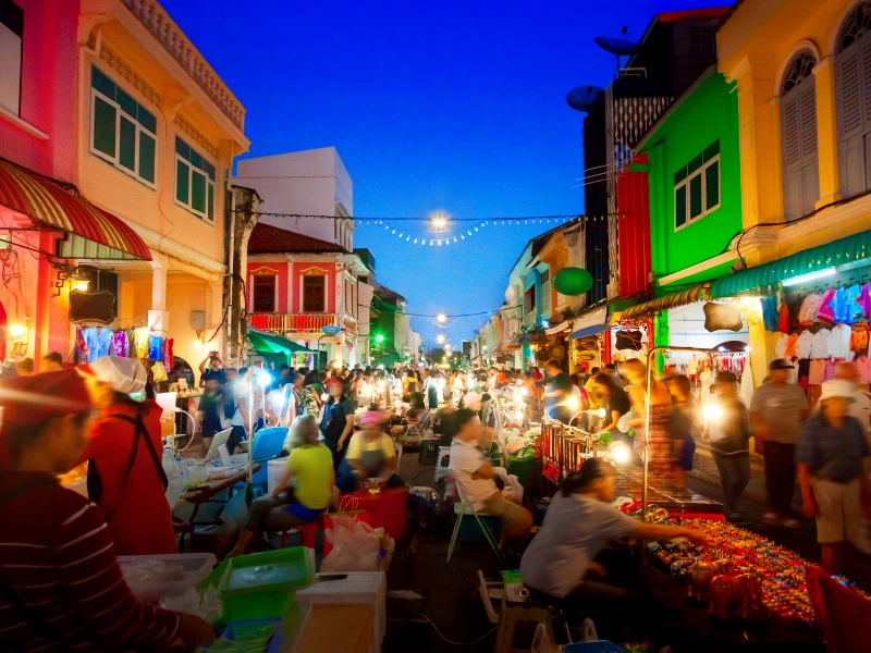 Evening street scene in Phuket Old Town with colorful buildings, market stalls, string lights, and people shopping under a deep blue sky.
