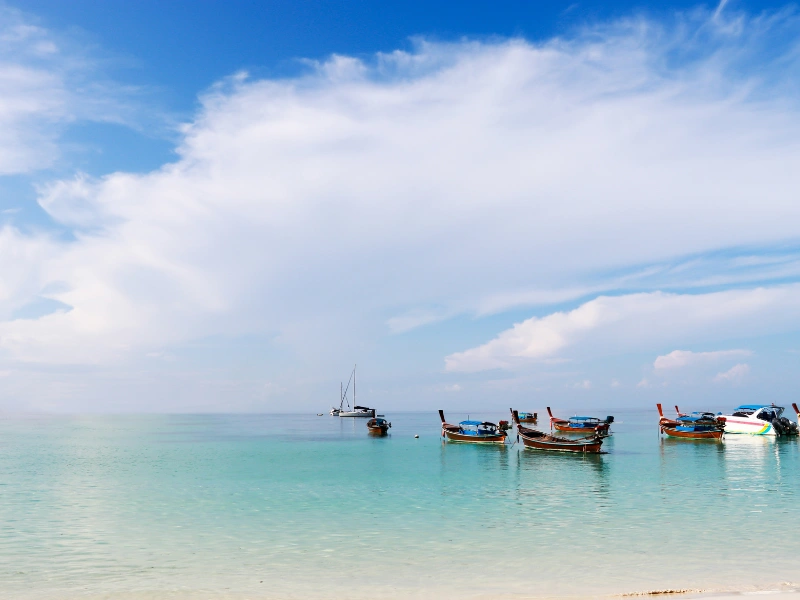 A serene beach with turquoise water, anchored boats near the shore, and a distant sailboat under a mostly clear sky.