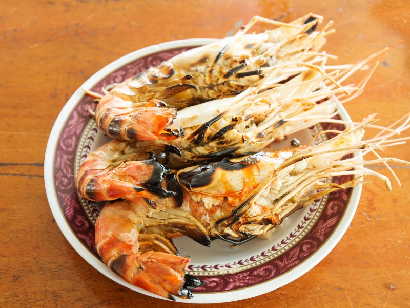 A plate with five grilled prawns placed on it. The prawns have a charred appearance, indicating they have been cooked over an open flame or grill. The plate has a decorative pattern around the rim and is set on a wooden surface.