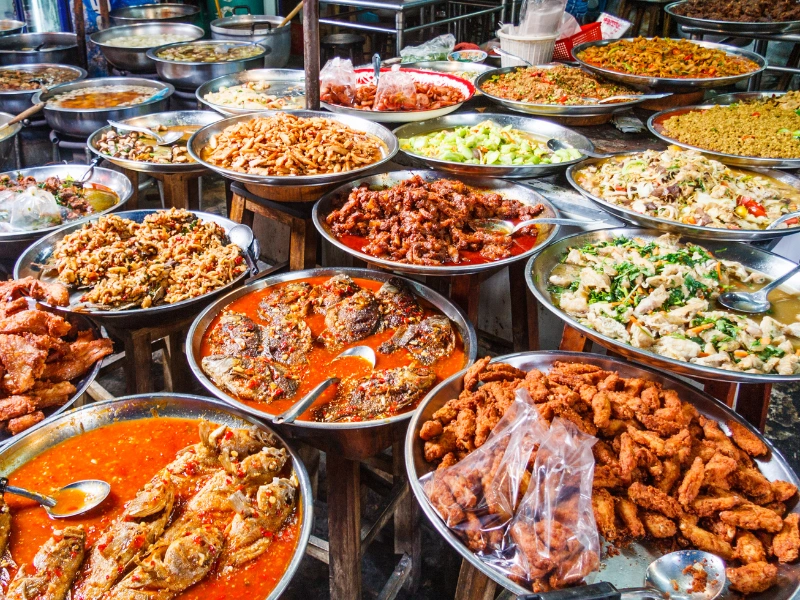 The image shows a variety of dishes displayed in large metal bowls at what appears to be a food market or street food stall. The dishes include various types of curries, fried foods, and other prepared meals.