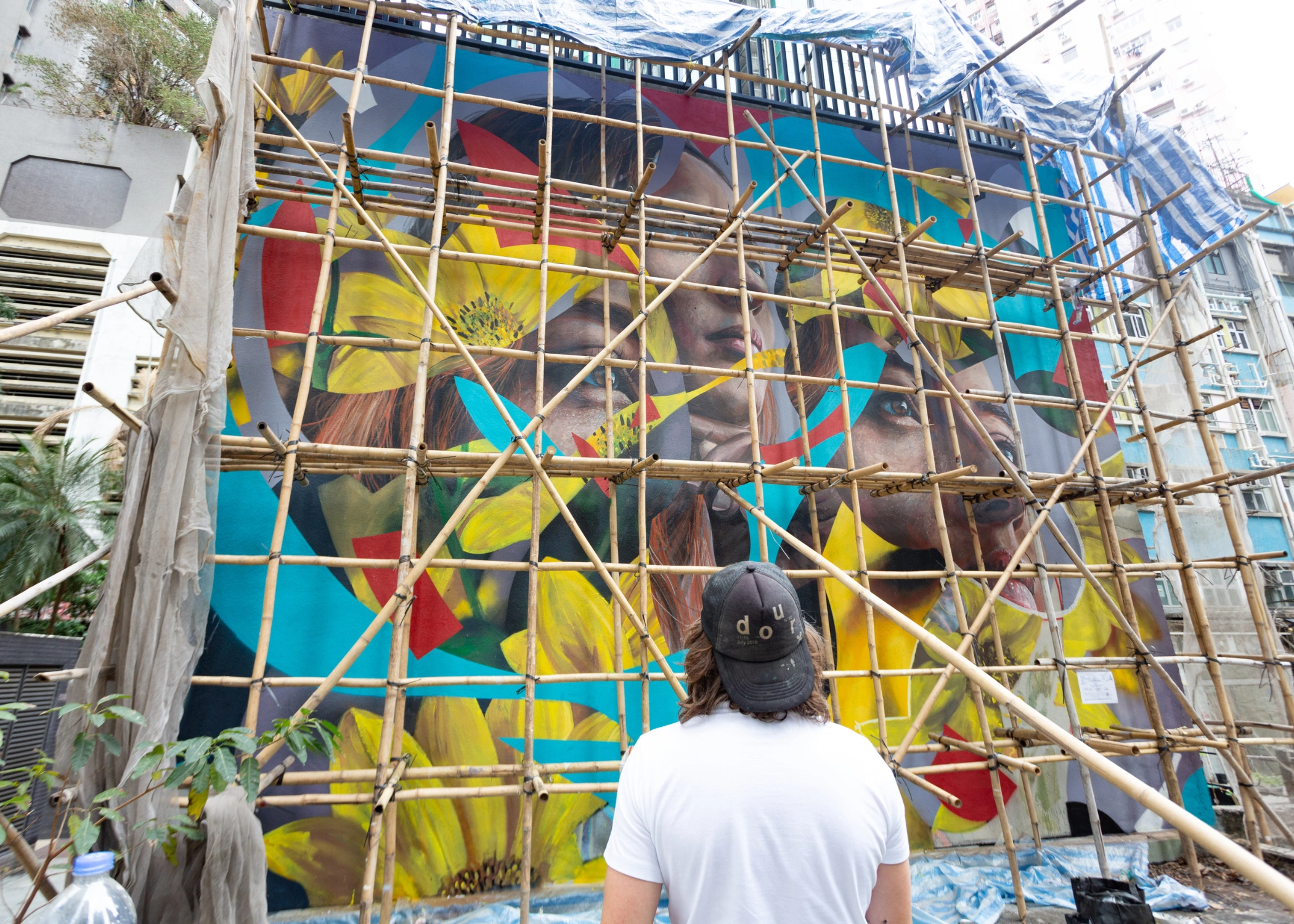Large mural on a building wall, partially covered by scaffolding, featuring vibrant faces and flowers; a person in a white shirt and black cap stands in front observing it.