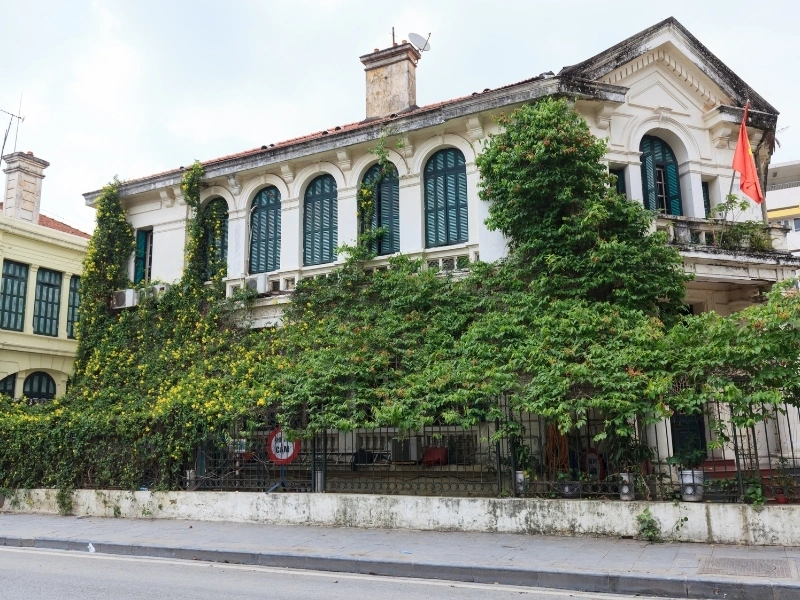 Two-story building with arched windows, red-tiled roof, and ivy-covered facade, featuring a small balcony and a red flag on the right side, on an empty street.