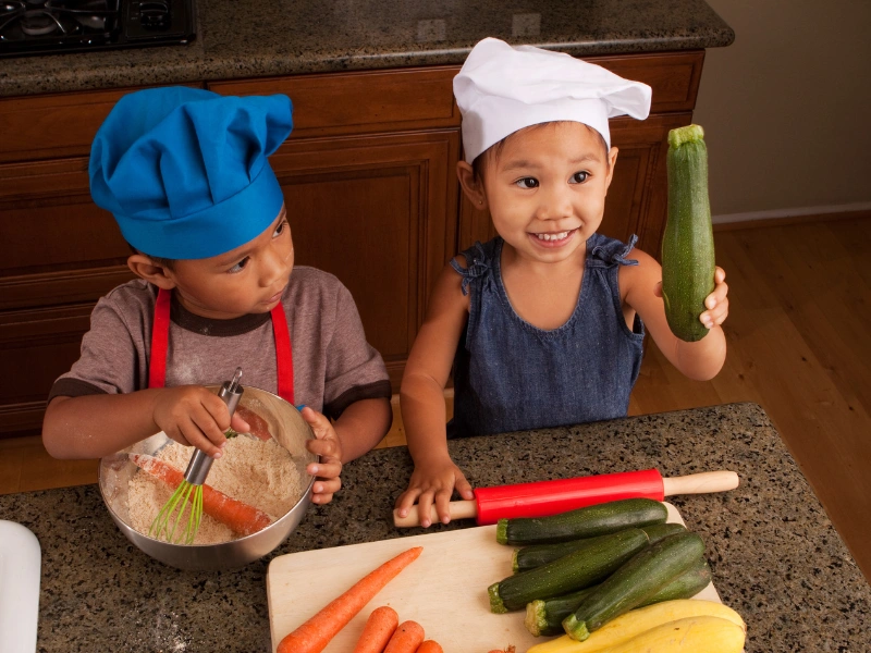 Two children in a kitchen, one mixing ingredient in a bowl while wearing a blue chef's hat and red apron, and the other holding a zucchini with a white chef's hat. Various vegetables are on the counter.