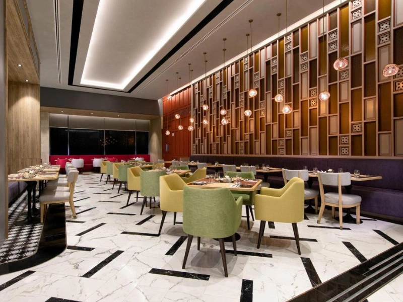 Modern restaurant dining area with green and yellow chairs around wooden tables, white marble flooring with black accents, and warm lighting—designed to be inviting and suitable for families with children.