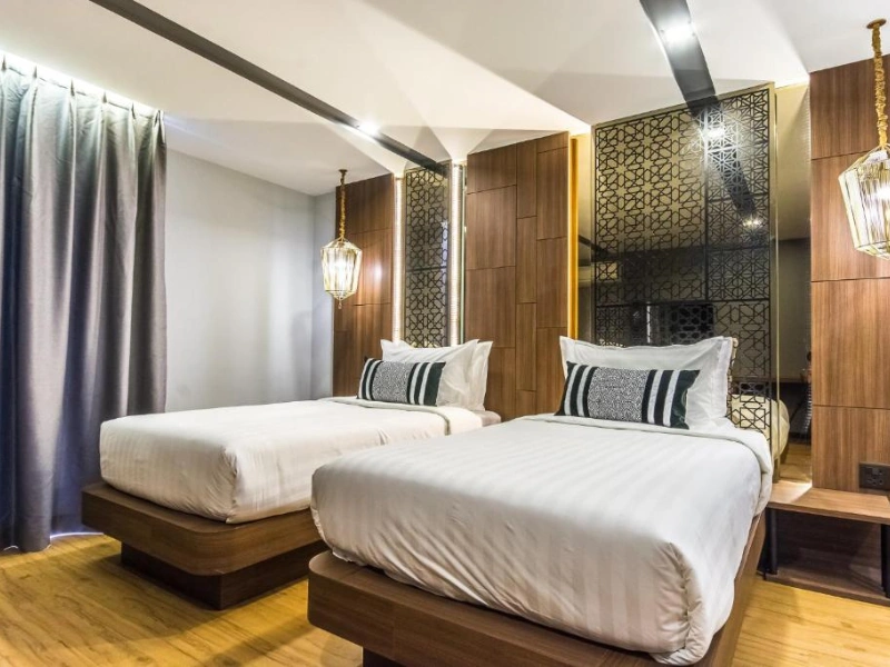 Modern hotel room in Nimman, Chiang Mai, featuring two single beds with white linens and striped pillows, wooden paneling, decorative hanging lights, a patterned mirror, and grey curtains.