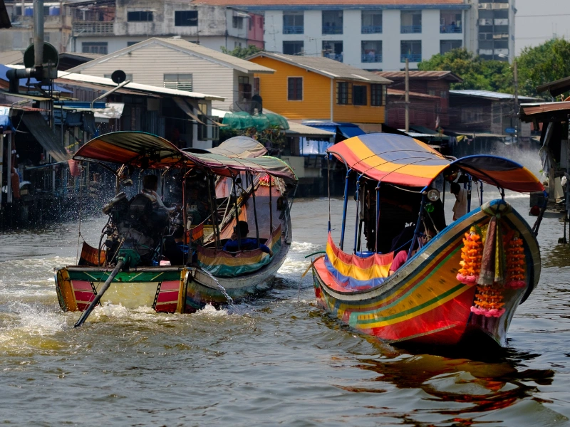 Two colorful long-tail boats travel through a canal lined with houses and buildings. One boat creates splashes with its engine, while the other is decorated with bright garlands.