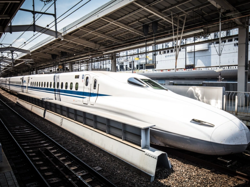 A sleek white Shinkansen bullet train with blue accents parked at a covered station platform, with overhead wires and infrastructure visible in the background.