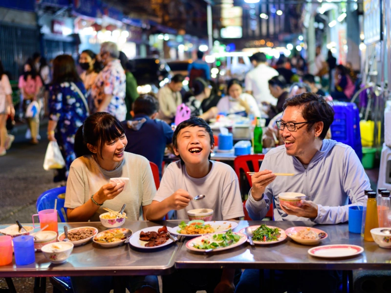 Four people sitting at an outdoor table enjoying a meal with various dishes, set in a busy street market or food court with blurred faces for privacy.