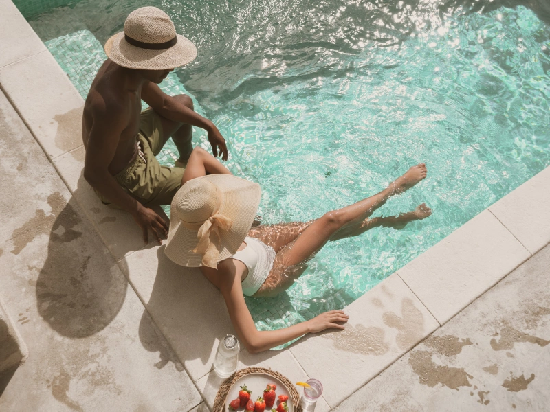The image shows two people sitting by the edge of a swimming pool. One person is wearing a hat and green shorts, while the other person is wearing a hat and a white swimsuit. The person in the white swimsuit has their legs in the water. There is also a plate with fruit and two drinks placed on the poolside near them.