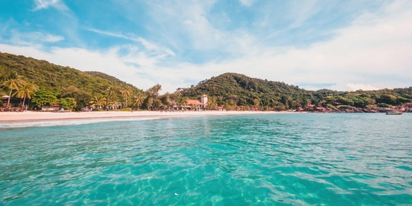 Clear turquoise sea water with green forested hills and a white sand beach under a bright blue sky at Redang Island