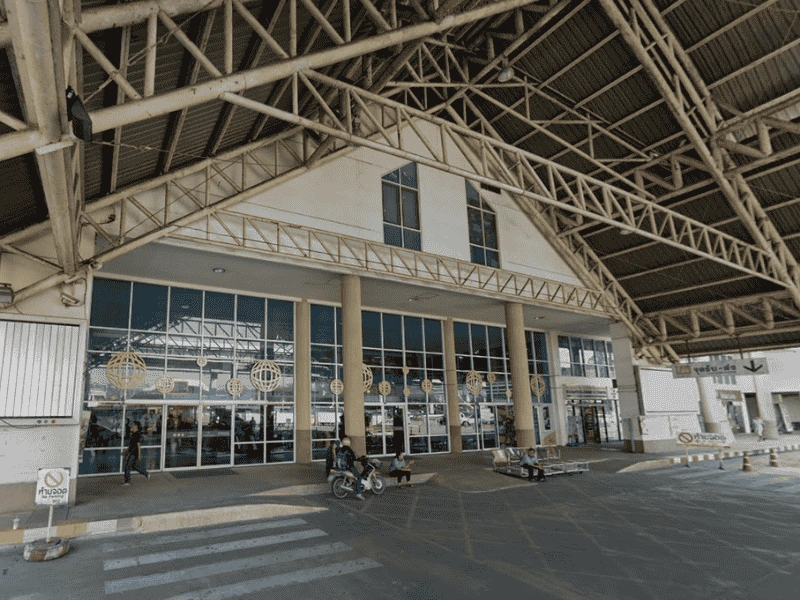 The exterior of a building with a large, triangular roof supported by metal beams. The building has a glass facade with multiple windows and doors. There are three columns at the entrance, and several people can be seen near the entrance, including someone on a motorcycle and others walking or standing. There is also a zebra crossing in front of the building.