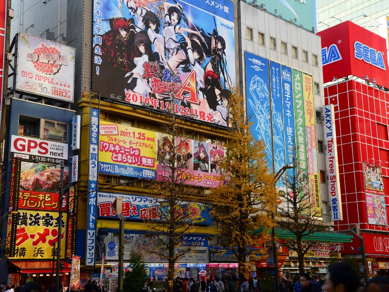 Vibrant street scene in Akihabara, Tokyo, with buildings covered in colorful anime advertisements, a prominent Sega building, and people walking along tree-lined sidewalks.