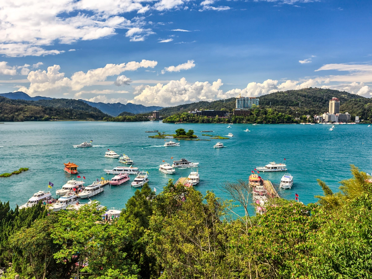 A scenic lake view with boats docked and sailing on the water, surrounded by lush green hills and distant mountains, with buildings including a tall hotel or resort visible across the lake under a partly cloudy sky.