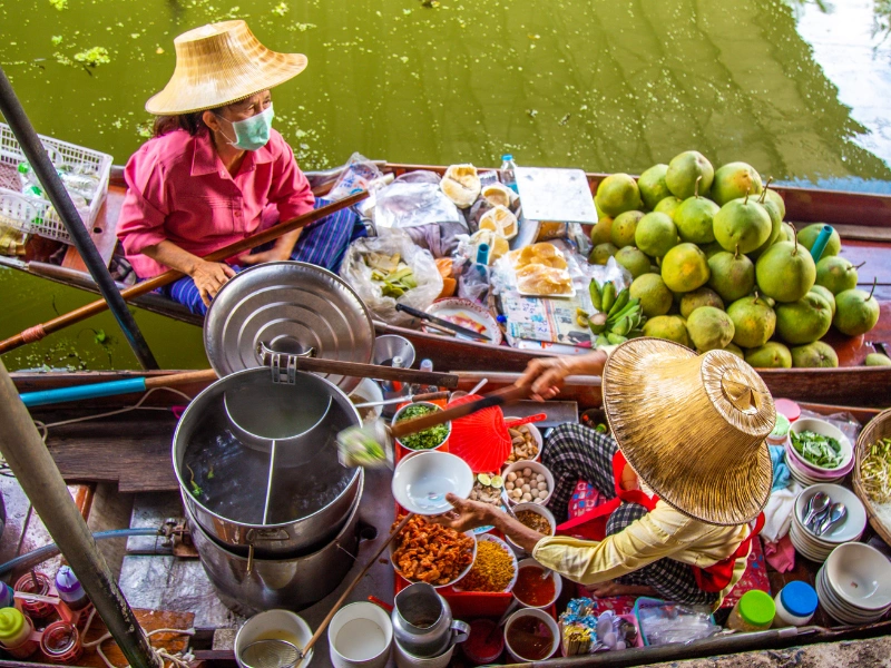 Two people in a boat on a body of water, likely a river or canal. The person on the left is wearing a pink shirt, blue pants, and a straw hat while sitting in the boat. The person on the right is wearing a yellow shirt, red apron, and straw hat while preparing food. There are various food items and cooking utensils around them, including coconuts, bowls of ingredients, and cooking pots.