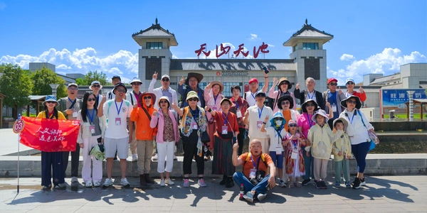 The sky is brilliant blue with dramatic white clouds. Happy tour group holding a red travel banner in front of the Tianshan Heavenly Lake entrance gate.