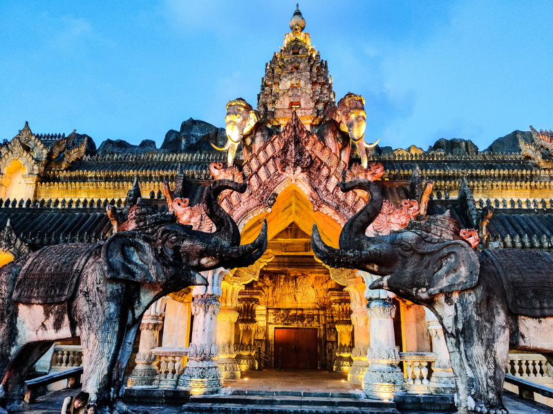 Ornate temple entrance with intricate carvings, flanked by two large elephant statues, illuminated under a dusk sky.
