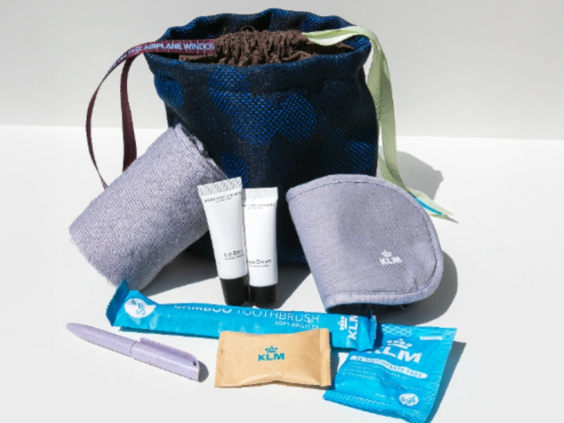 A KLM-branded airline amenity kit displayed on a white surface. The kit includes a dark blue patterned pouch with ribbon ties, a grey eye mask, a pair of socks, two small tubes of skincare products, a toothbrush in blue packaging, toothpaste, and a purple pen-like item.