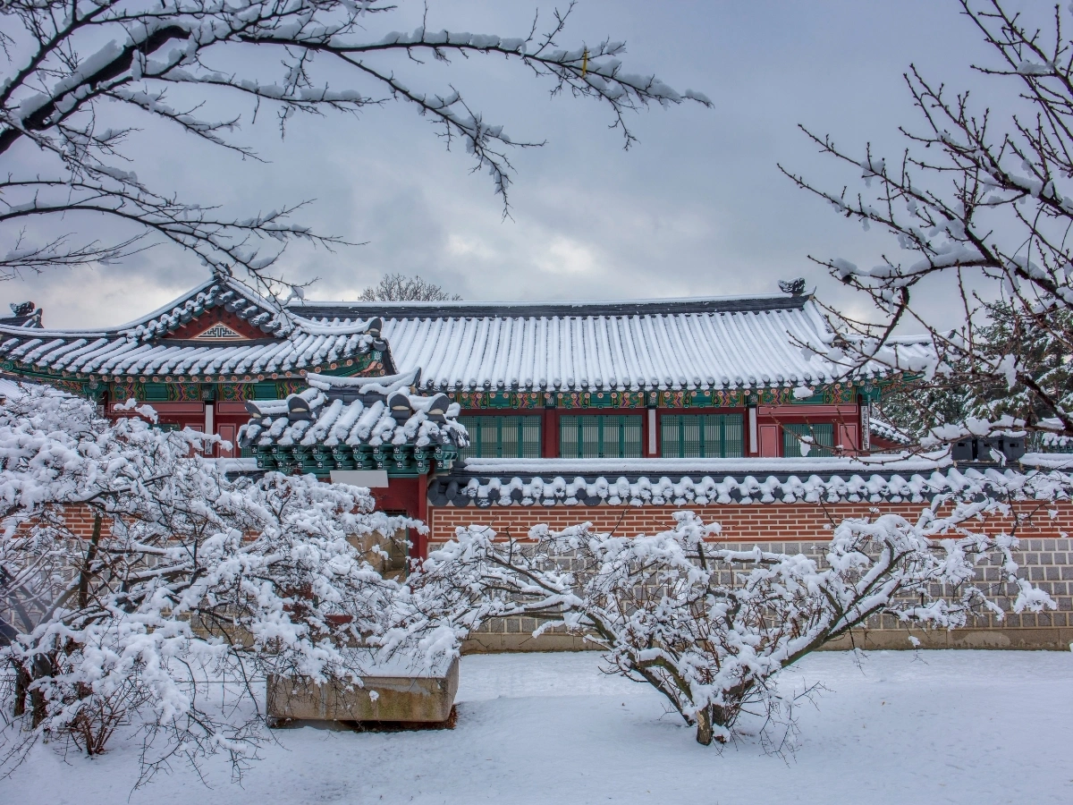 Traditional Korean building with a snow-covered tiled roof, red and white walls, and green windows, set in a winter landscape with snow blanketing the ground and tree branches. The architecture showcases intricate Korean design elements under an overcast sky