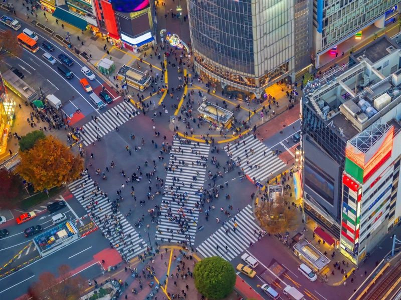 Aerial view of Shibuya Crossing in Tokyo, Japan, with large crowds of pedestrians crossing in multiple directions, surrounded by tall buildings and bright advertisements.