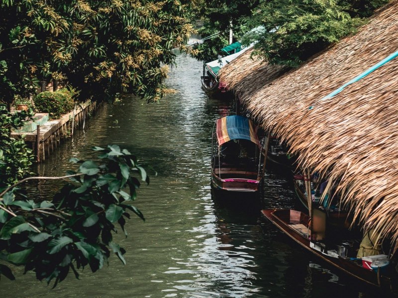 A narrow waterway or canal with boats docked along the sides. There are trees and greenery on both sides of the canal, and a thatched-roof structure is visible on the right side. The water appears calm, and there are no people visible in the image.