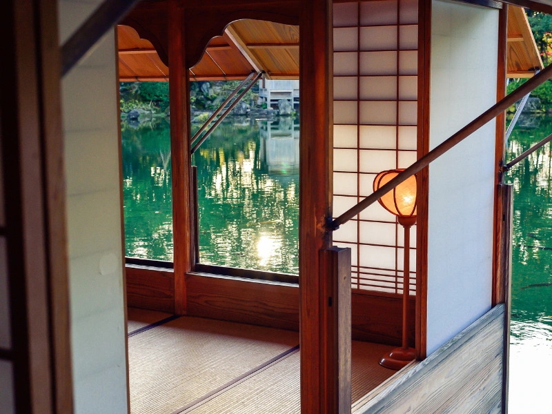 Traditional Japanese tatami room with sliding shoji doors, tatami mats, wooden beams, and a paper lantern, overlooking a calm body of water with tree reflections.