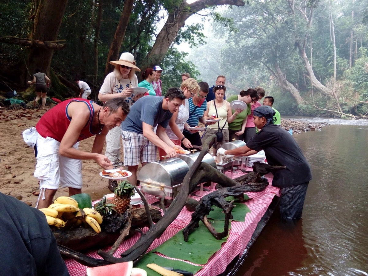 A group of people serving themselves from a buffet-style food setup on a red and white checkered tablecloth beside a river in a forested area, with bananas, pineapples, and other dishes surrounded by dense greenery.