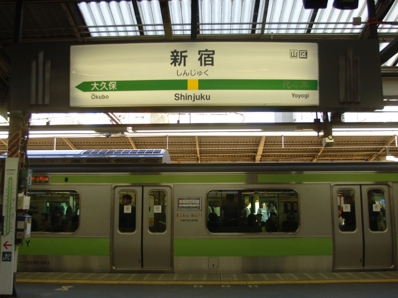 Train at Shinjuku Station platform with a bilingual sign displaying 'Shinjuku' in English and Japanese, along with directions to Okubo and Yoyogi stations.