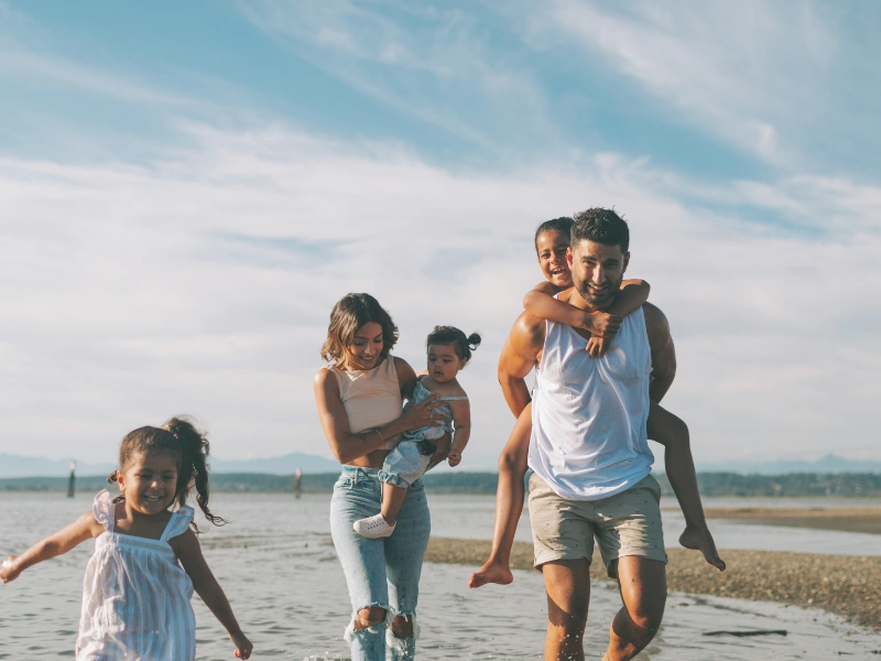 The image shows a family of four walking along a beach. The adults are carrying the children, with one adult holding a child in their arms and the other giving a piggyback ride. The family is dressed in casual summer clothing, and the background features a clear sky with some clouds and distant mountains.