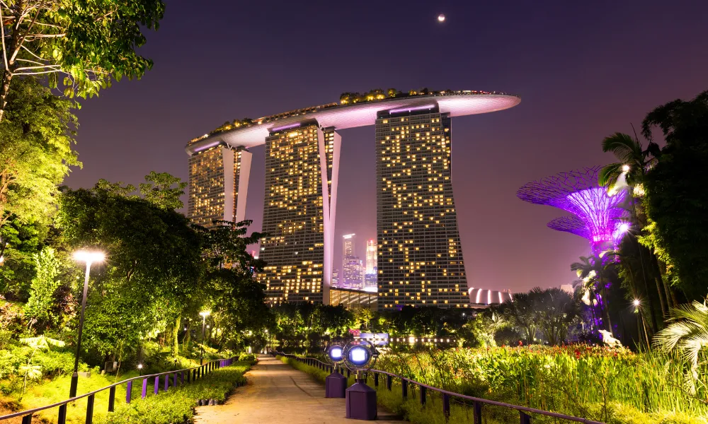 A nighttime view of Marina Bay Sands in Singapore, featuring its three illuminated towers connected by a sky park on top. The surrounding area is lush with greenery and well-lit pathways, with the Supertree Grove visible to the right, glowing in purple light.