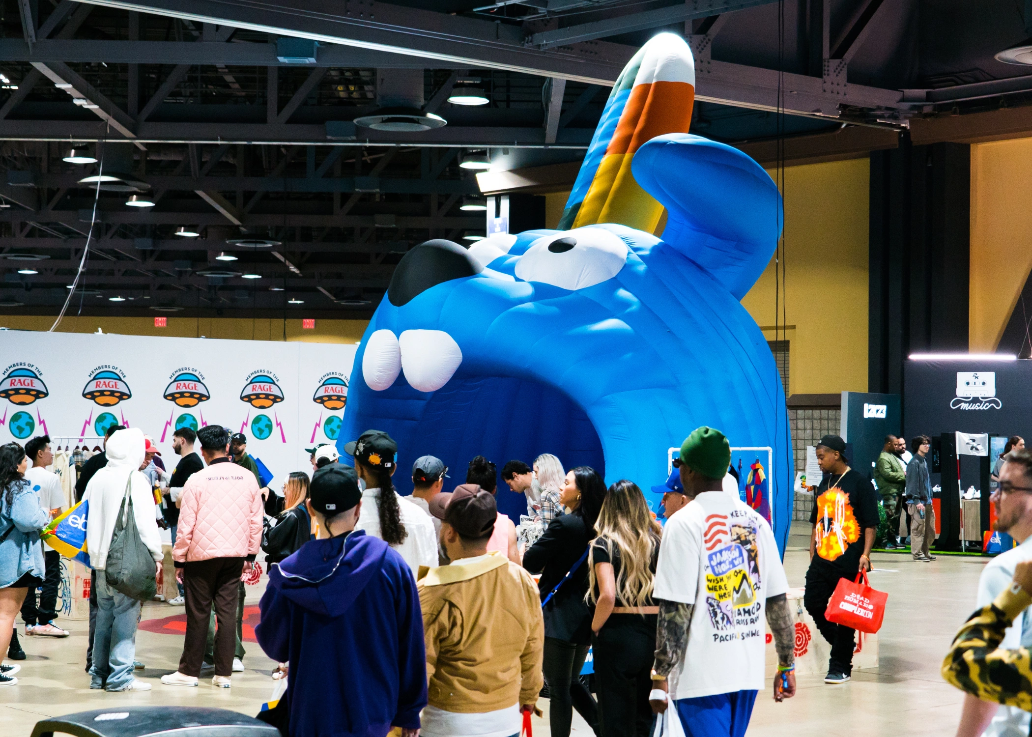 Large indoor event with crowds gathered around a giant inflatable blue cartoon character with an open mouth, big teeth, and colorful ears; booths and displays featuring the same character are visible in the background.