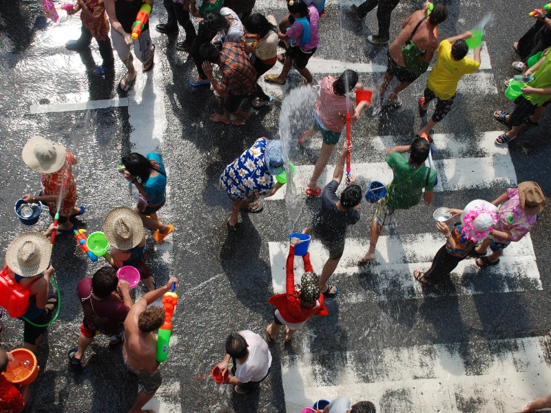 A lively crowd celebrating Songkran festival in Thailand, engaging in water fights with buckets, water guns, and hoses on a wet street. Participants wear colorful clothes and hats, with water splashing everywhere.