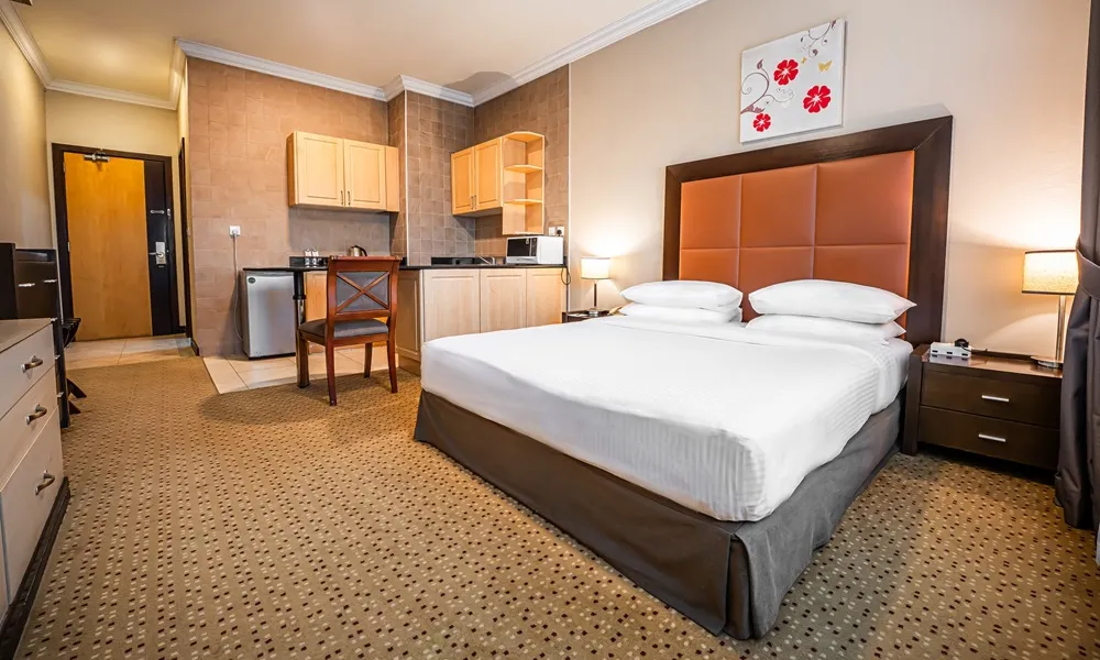 Spacious guest room at Kingsgate Hotel Doha featuring a queen-size bed with white linens, modern headboard, kitchenette, dining area, and warm neutral decor